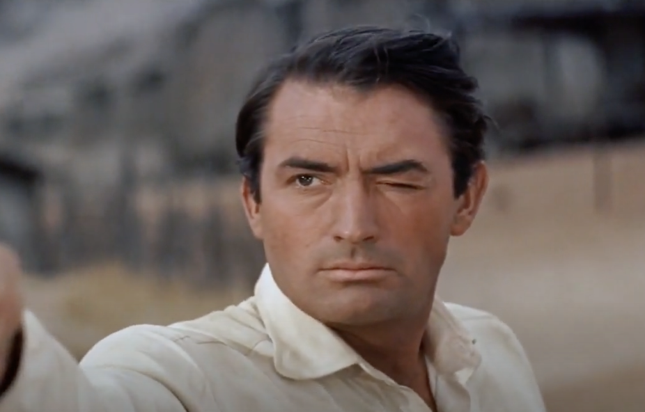 A man with short dark hair and a white shirt winks while pointing forward. The background is blurred, showing what appears to be a desert landscape and structures. His expression is focused and determined.