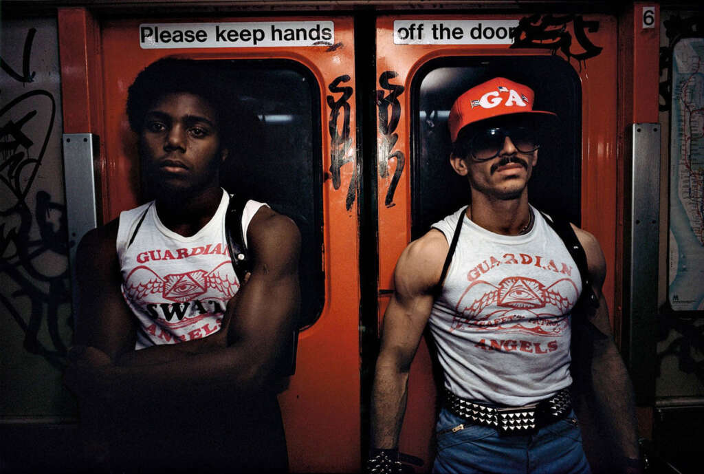 Two men in matching "Guardian Angels" shirts ride a graffiti-covered subway. One wears a red cap and sunglasses, the other has a backpack. A sign above them reads, "Please keep hands off the door.