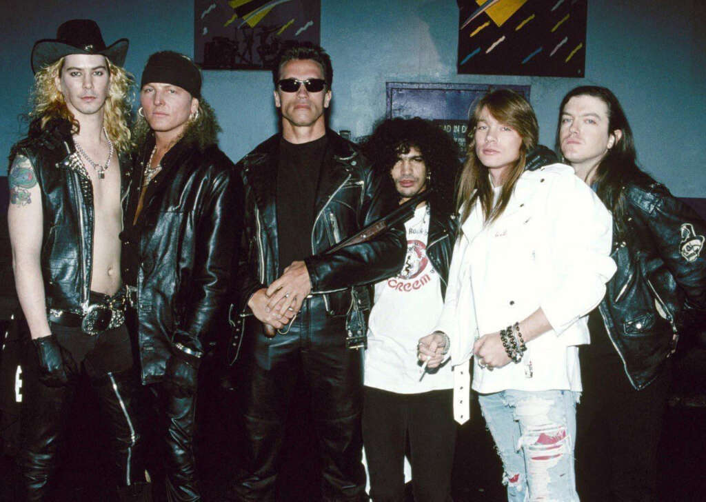 Six men stand in a group. Most are wearing leather jackets, and one is in a white jacket. They have various hairstyles, including long and curly, and some wear sunglasses. Behind them is a dark background with abstract art.