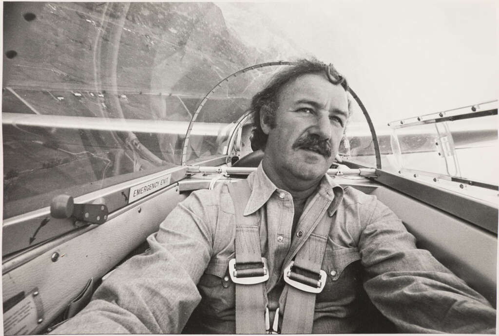 A man with a mustache is piloting a glider, wearing a harness. The black and white photo captures him in mid-flight, with the landscape visible through the canopy. Text reading “EMERGENCY EXIT” is visible on the cockpit's side.