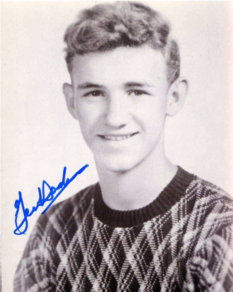 Black-and-white portrait of a young man with curly hair, smiling and wearing a patterned sweater. There is a blue signature overlaid on the image.