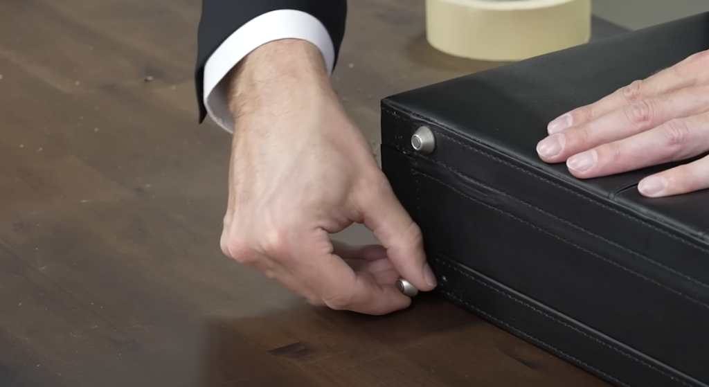 A person fastening a button on a black leather briefcase on a wooden table. They are wearing a dark suit with a white cuff. Nearby, there is a roll of beige tape.