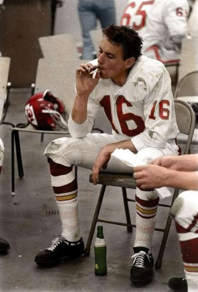 Football player in uniform with number 16 sits on a folding chair in a locker room, smoking a cigarette. A green bottle is on the floor nearby, with teammates and equipment in the background.