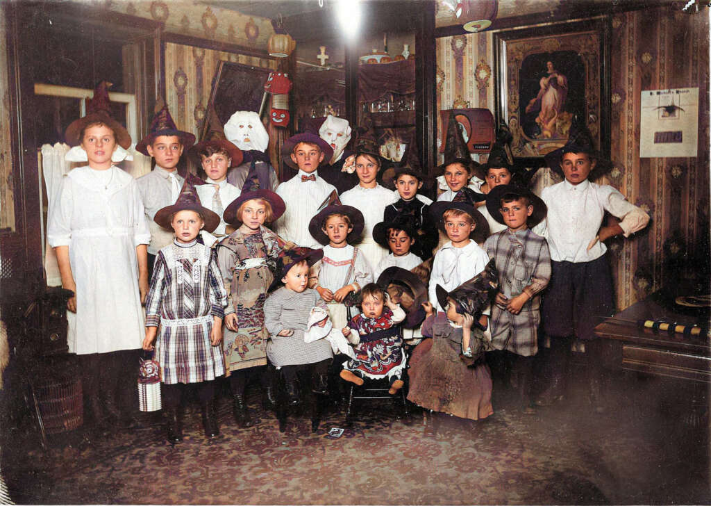 A group of children dressed in early 20th-century clothing and various costumes, including hats. They pose indoors, surrounded by vintage decorations and a wall with framed pictures and patterns.