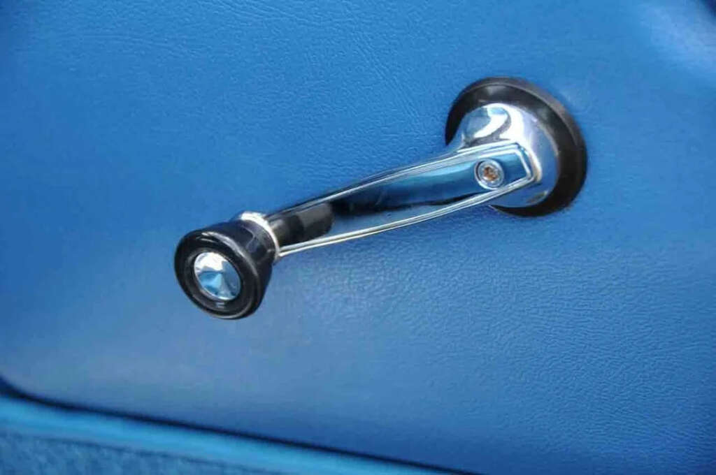 Close-up of a manual car window crank handle on a blue car door panel. The handle is shiny chrome with a black knob at the end, attached to the door, used for rolling windows up or down.