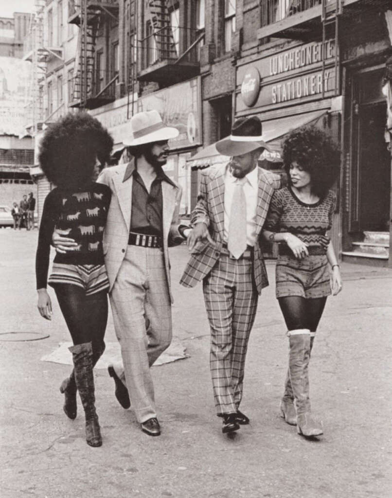 Four people walk down an urban street, dressed in 1970s fashion. Two men in wide-brimmed hats and suits have their arms linked with two women in stylish outfits and boots. They are smiling and chatting amidst a backdrop of storefronts.