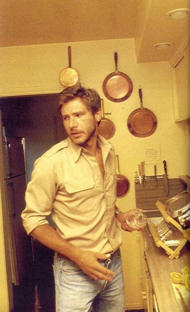 A man in a casual beige shirt and jeans stands in a small kitchen, holding a glass. Copper pans hang on the walls, and there are dishes drying on a rack on the counter. The lighting is warm and dim.