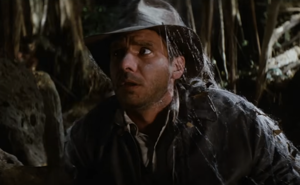 A man in a weathered hat and jacket, covered in cobwebs, looks concerned as he moves cautiously through a dim, jungle-like environment. Vegetation and shadows surround him, contributing to the scene's tense atmosphere.