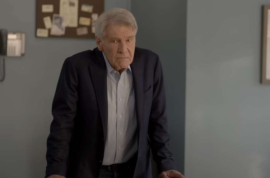 An older man with gray hair, wearing a navy blazer and light blue shirt, leans on a table in an office. The wall behind him features a bulletin board with various papers and a light switch.