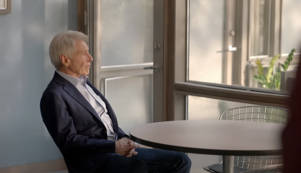 An older man with gray hair sits in a chair at a round table, looking out a window. He is wearing a dark blazer, light shirt, and jeans. The room is softly lit and the window offers a view of greenery outside.