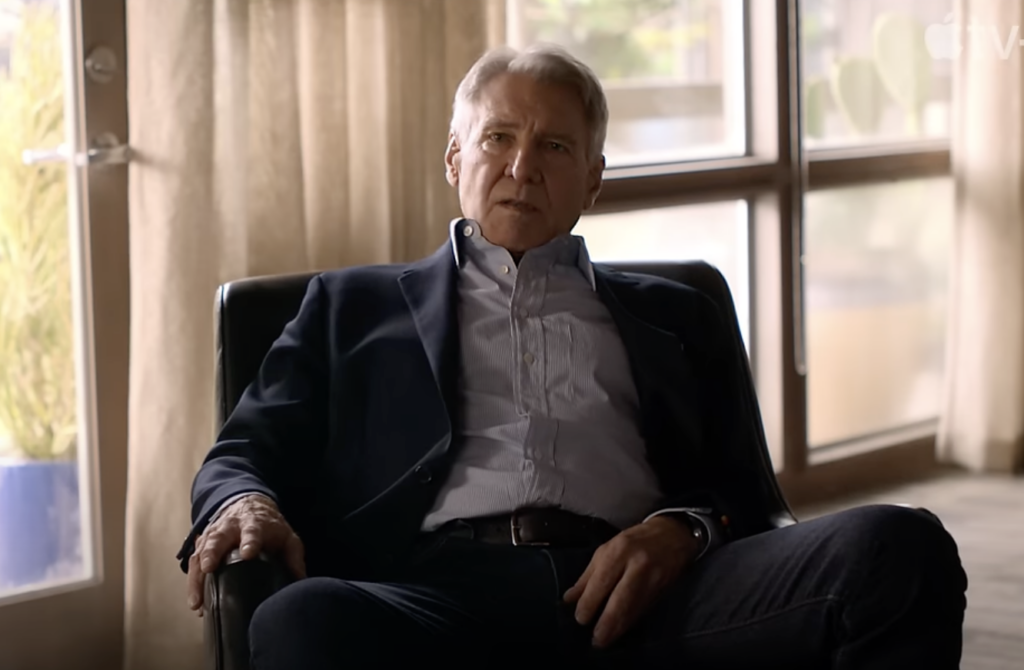 A man with short gray hair is seated in a black chair, wearing a dark blazer, light-colored shirt, and jeans. He is in a room with large windows and light curtains, and he appears to be engaged in a conversation.