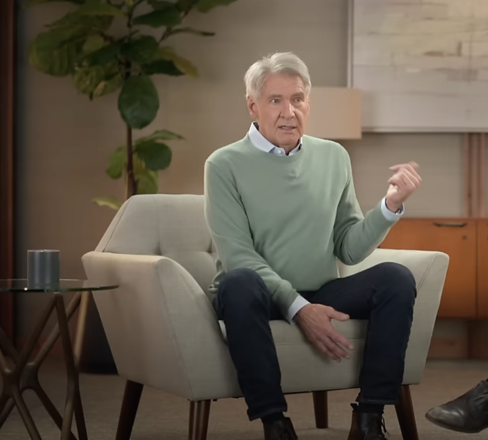 An older man with gray hair sits in a light gray armchair, wearing a light green sweater and dark jeans. He gestures with his hand while talking. There's a potted plant and framed artwork in the background. A small glass table is beside him.