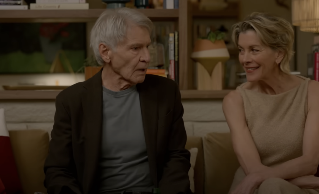 An older man and woman sit on a sofa in a cozy living room, engaged in conversation. The man has gray hair and wears a dark jacket over a shirt, while the woman has short blonde hair and wears a light-colored sleeveless top, smiling warmly.