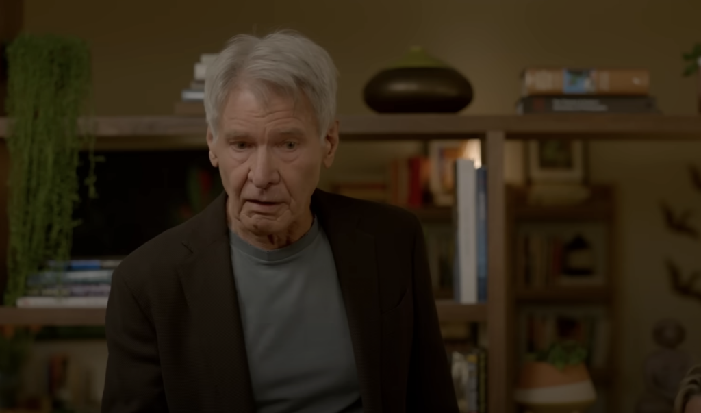 An older man with gray hair stands indoors, wearing a dark suit jacket over a gray shirt. Behind him is a wooden shelf filled with books and plants, and the room is warmly lit.