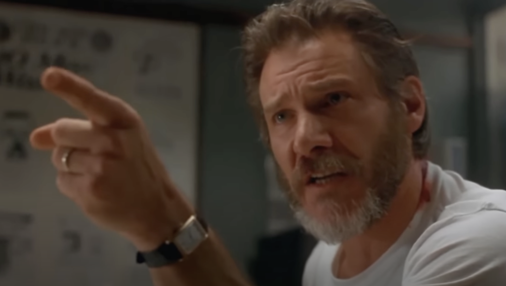 A man with a beard and mustache is pointing forward and appears to be speaking assertively. He is wearing a white shirt and a watch on his left wrist. Behind him is a wall with various documents and charts.