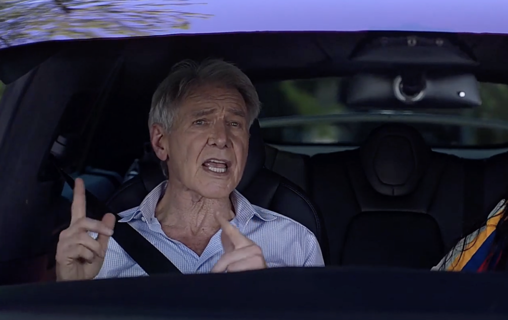 An older person with gray hair is sitting in the driver's seat of a car, wearing a seatbelt. They appear to be talking or explaining something, with both hands slightly raised. The interior of the car is dark.