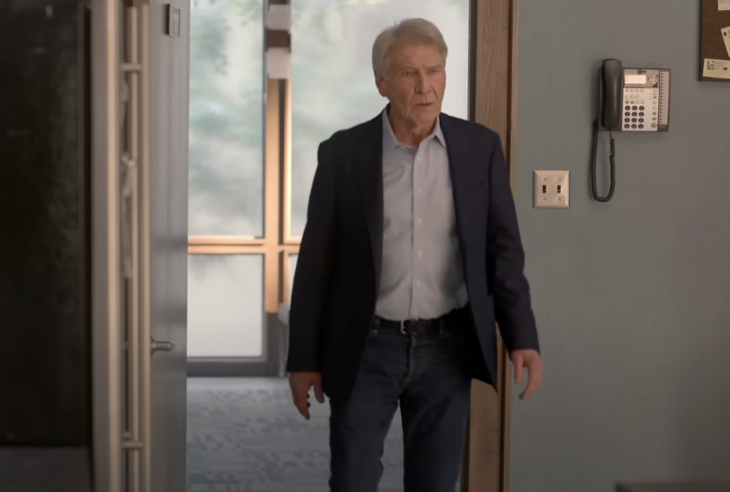 A person with gray hair is walking through an open doorway into a room. They are wearing a dark jacket over a light shirt and jeans. The room has a wall-mounted phone and a corkboard. Sunlight is streaming through large windows in the background.