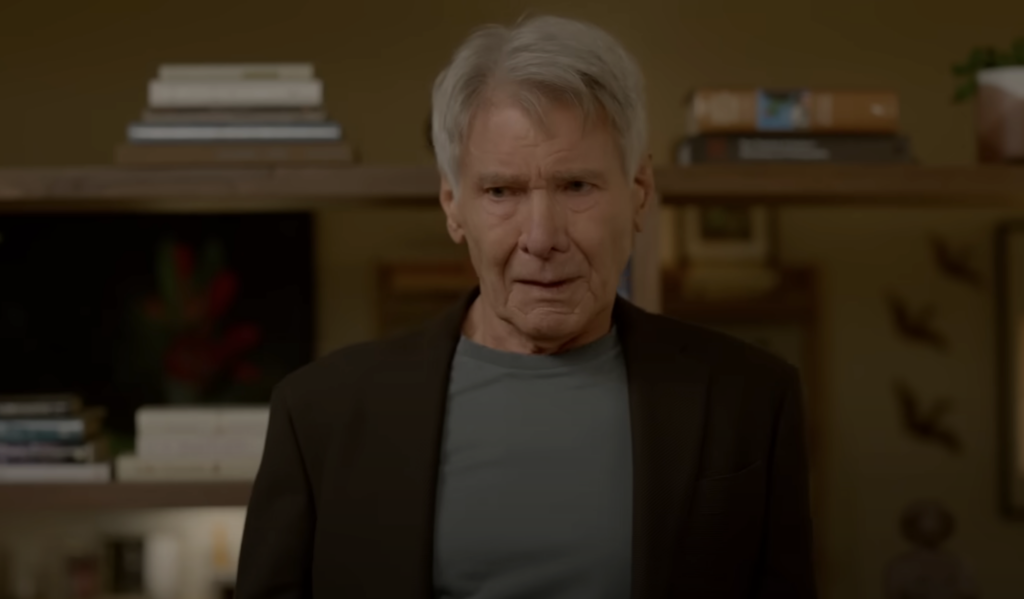 An older man with gray hair stands indoors, wearing a dark blazer over a gray shirt. He appears to be concerned or deep in thought. Books and a plant are visible in the background on a shelf.