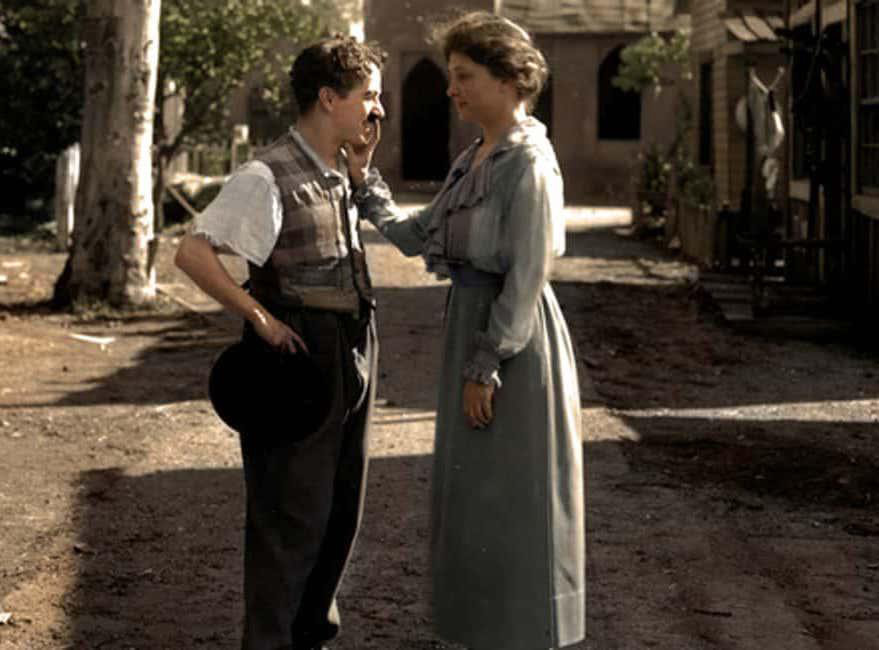 A woman in a long dress tenderly touches a man’s face with her hand. The man, holding a hat, smiles at her. They're standing outside on a dirt path, with trees and buildings in the background. The scene is in a nostalgic, sepia-toned color.