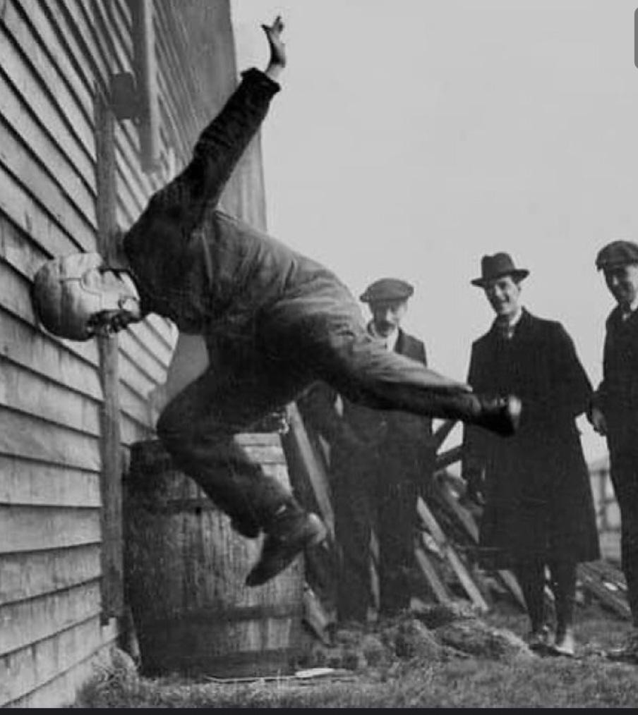 A man in protective headgear crashes sideways into a wooden wall, demonstrating a stunt. Three men in early 20th-century attire watch from a distance. A barrel and scattered hay are in the background.