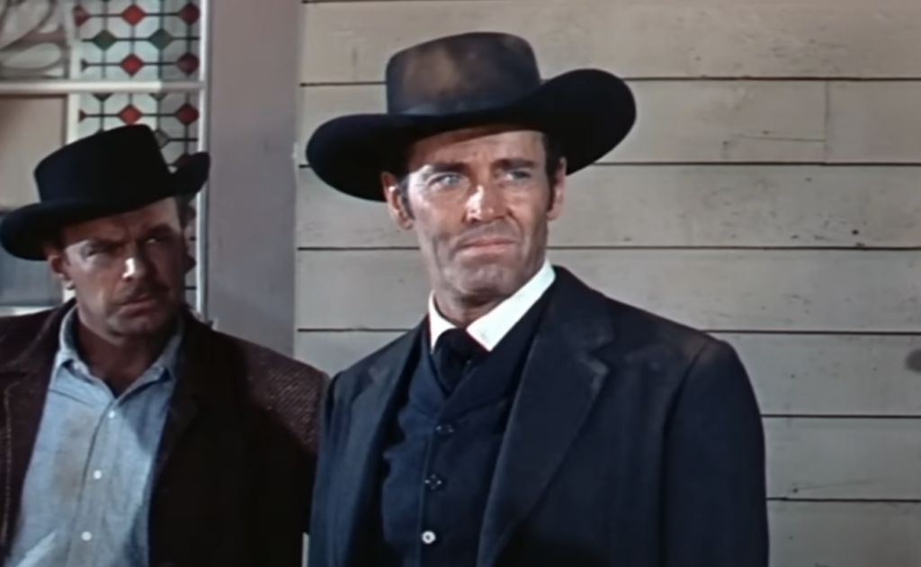 Two men in Western attire stand against a wooden wall. The man in front wears a black hat, suit, and vest, looking serious. The man behind, also in a hat, gazes off to the side. A hint of a patterned window appears in the background.