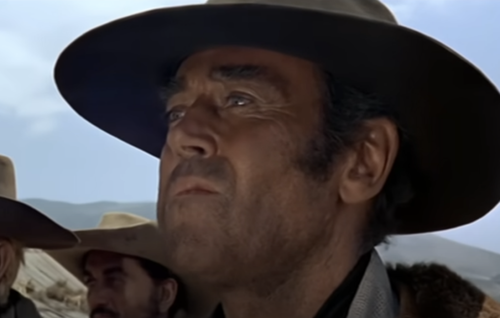 A close-up of a man with a serious expression, wearing a wide-brimmed hat. The background shows clear skies and blurred figures of other people also in hats.