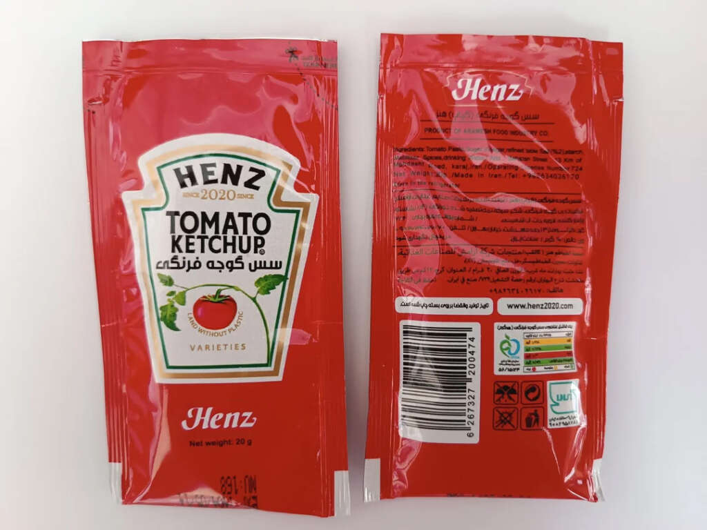 Two red packets of HenZ Tomato Ketchup, one showing the front with branding and a tomato image, and the other showing the back with text and certifications in multiple languages, including Arabic.