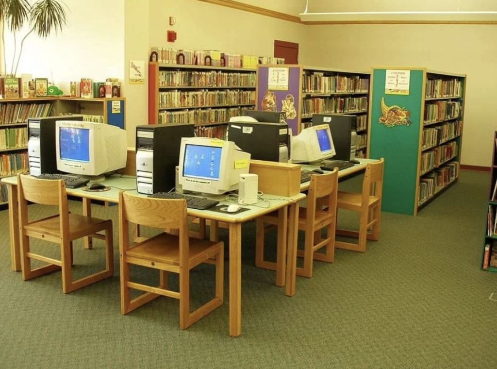 A library with bookshelves filled with books. In the foreground, there are several wooden tables with vintage computer monitors and chairs. The carpet is green, and the space is well-lit, creating a quiet study atmosphere.