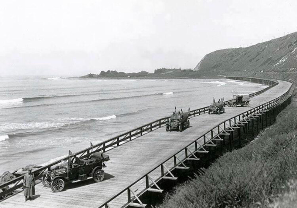 Vintage cars travel along a wooden coastal road with ocean waves on the left and a cliff on the right. A man stands near a car in the foreground, gazing at the scenery. The scene evokes a sense of early 20th-century travel.