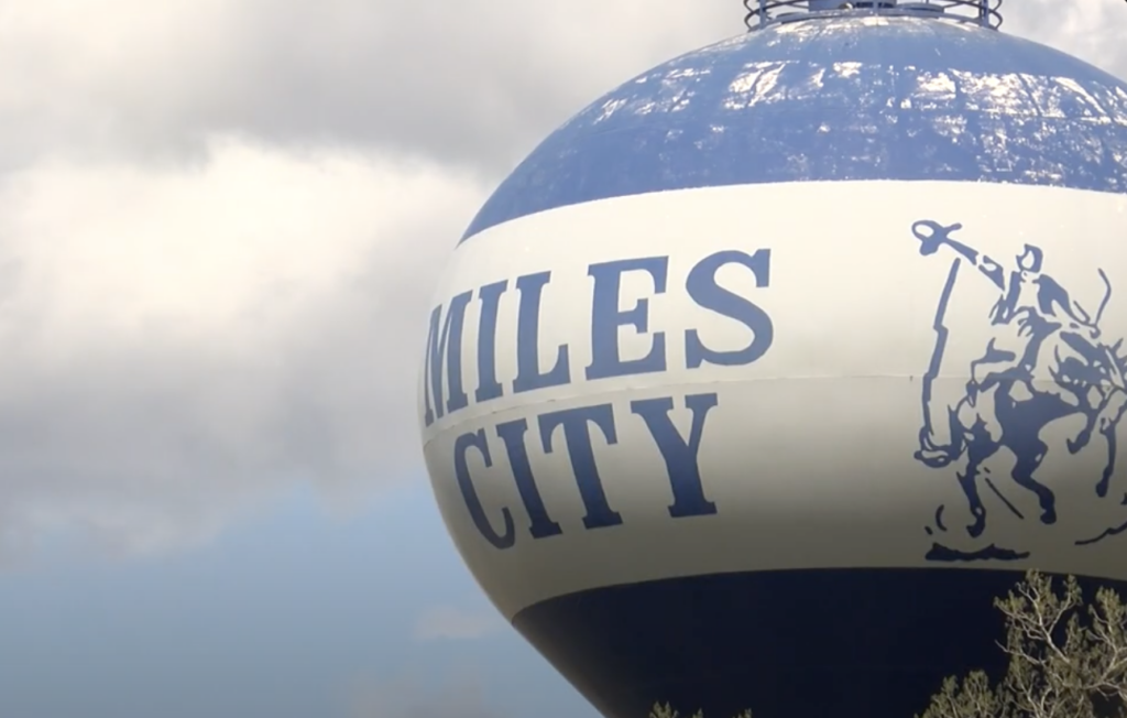 Water tower painted with "Miles City" and an image of a cowboy riding a horse. The tank is set against a cloudy sky background.