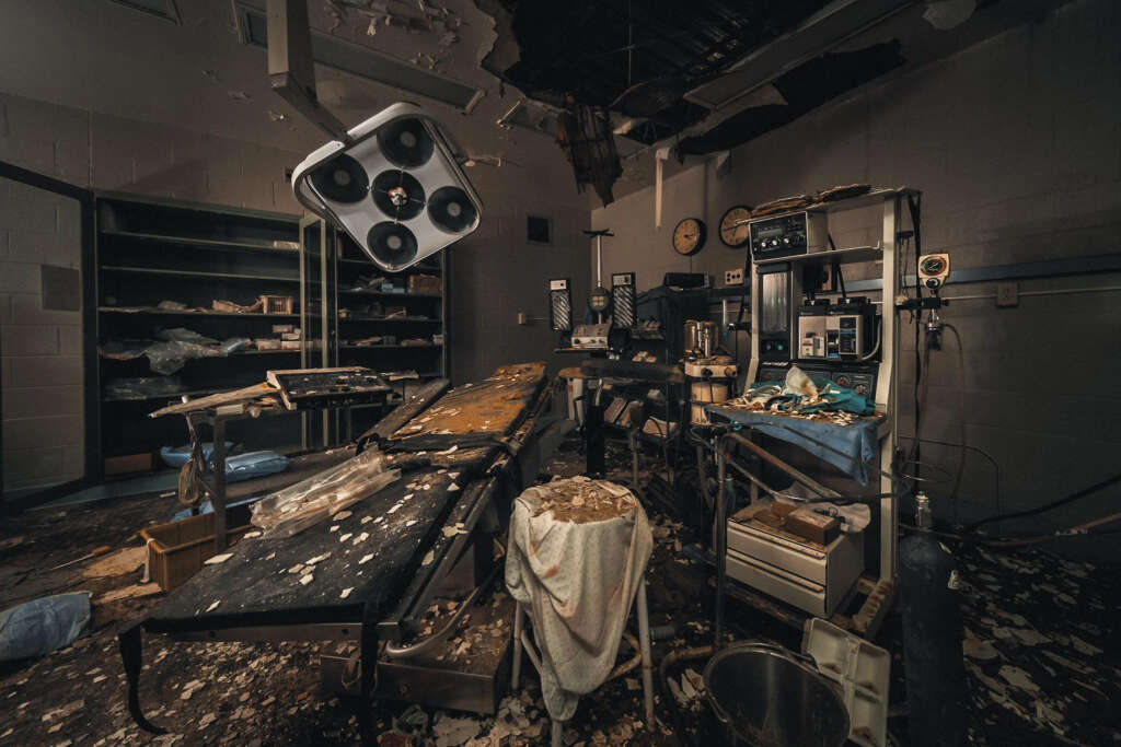 An abandoned operating room with decaying tables and equipment. The ceiling is damaged, and debris is scattered across the floor. Shelves are partially empty, and medical devices are old and unused. The room is dimly lit, creating an eerie atmosphere.