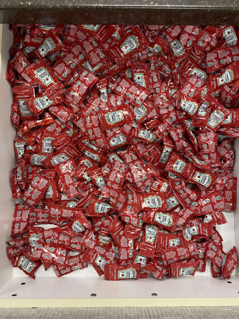 A large pile of red ketchup packets fills a drawer. Each packet is small and individually wrapped, with the brand label visible on some of them. The packets are stacked haphazardly, creating a dense and crowded appearance.