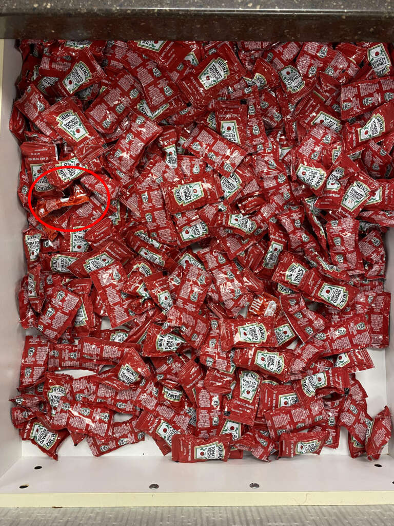 A large pile of red ketchup packets fills a container. Among the packets, one is circled in red. Each packet has a white label with black text and a logo.