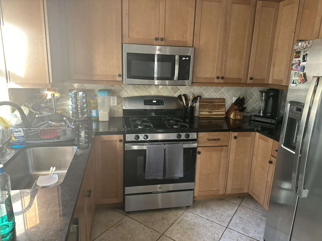 A kitchen with wooden cabinets features a stainless steel oven and microwave. A refrigerator is on the right, and a sink with dishes on a rack is on the left. Various kitchen items are on the countertops, with a diamond-patterned backsplash.