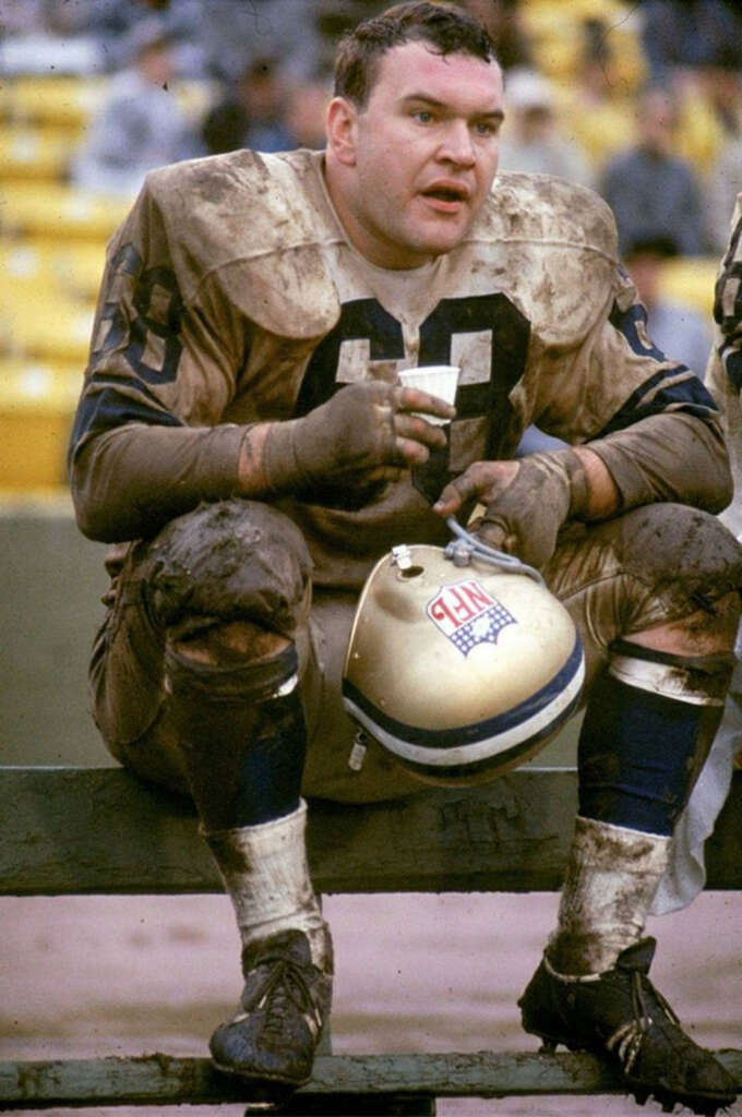 A football player wearing a muddy uniform sits on a bench, holding a cup in one hand and a helmet in the other, with a tired expression. The stadium seats in the background are mostly empty.