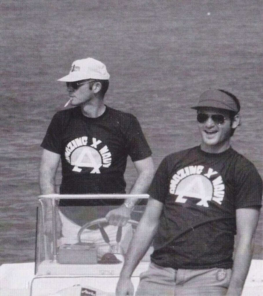 Two men on a boat wearing matching "Amazing Ivan" T-shirts. One man, wearing a cap, is standing and smiling. The other man, also wearing a cap and sunglasses, is in the background looking to the side. The scene is by the water.