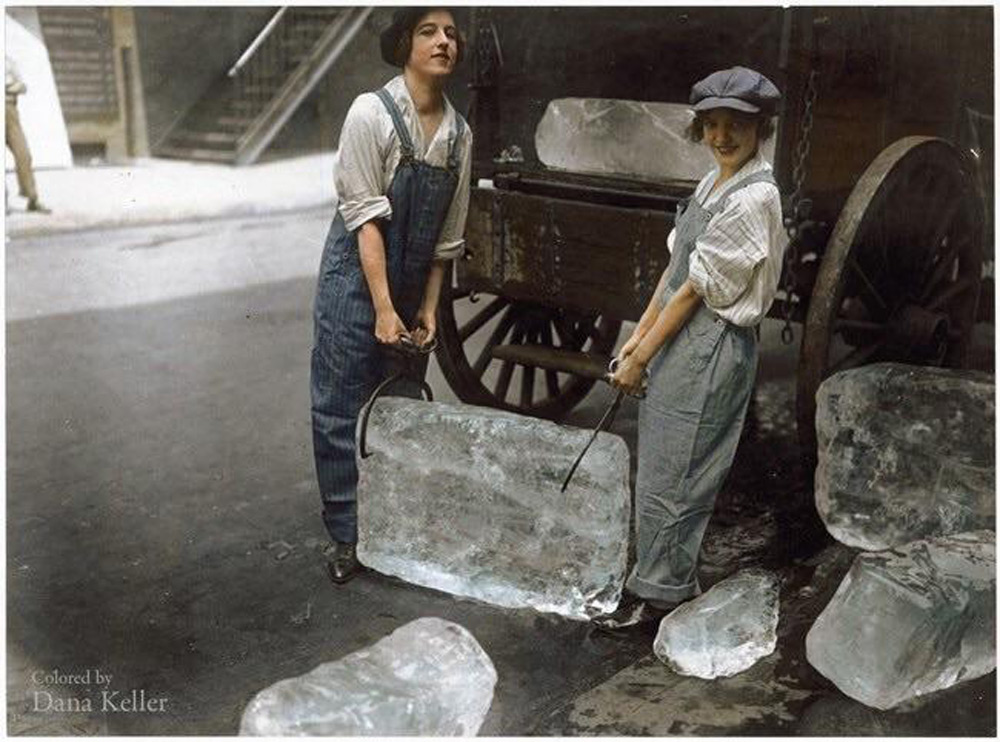 Two people in early 20th-century attire, wearing overalls and caps, are lifting a large block of ice with tongs. They stand beside a cart filled with more ice blocks on a street, with scattered ice chunks on the ground.