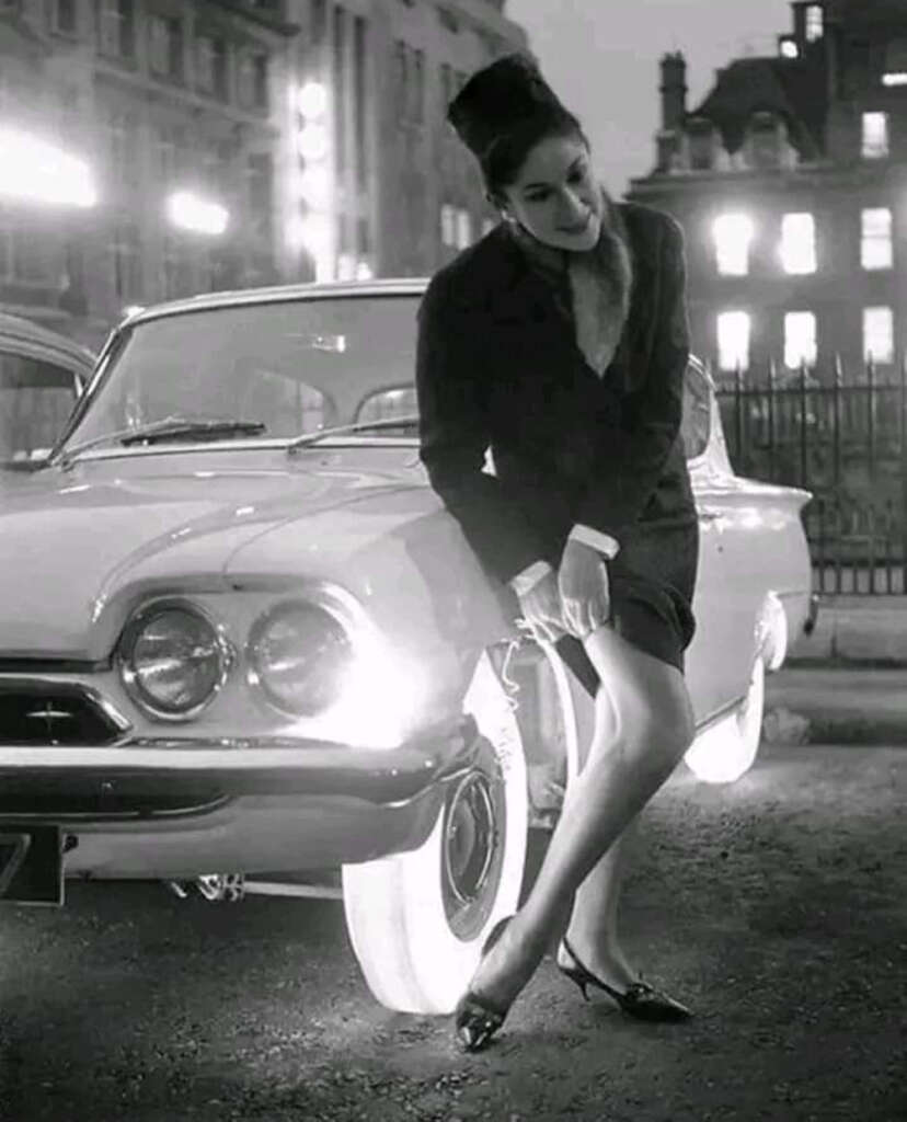 A woman in a vintage outfit leans against a classic car with illuminated whitewalls at night. The background features blurred buildings and lights, giving a retro urban feel.