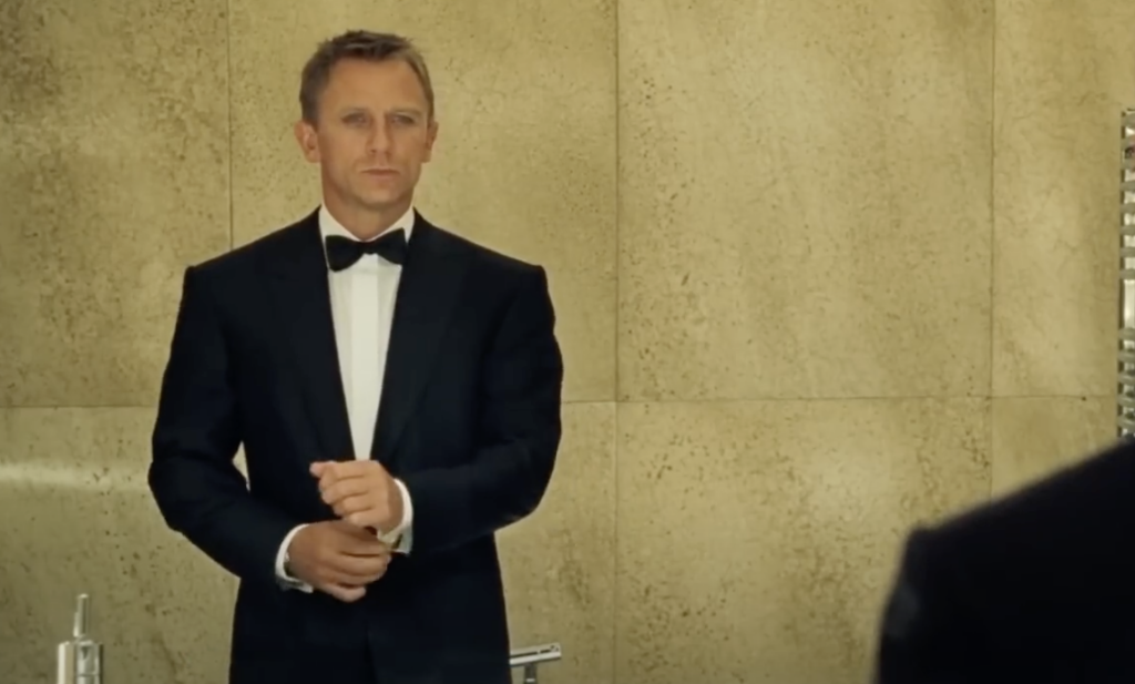 A man in a black tuxedo and bow tie stands in front of a mirror, adjusting his cufflink. The background features a beige, textured wall.