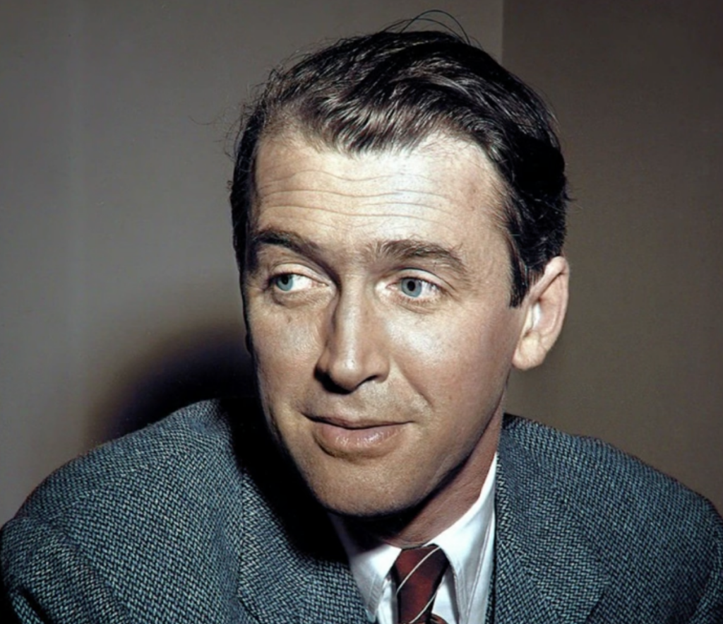 A man with combed-back hair and a suit gazes slightly to the side. He has a subtle smile and wears a patterned tie over a collared shirt. The background is a simple, muted tone.