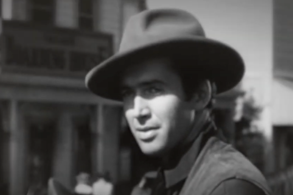 Black and white image of a man wearing a wide-brimmed hat and a vest, looking to the side. A building with blurred text is visible in the background. The scene has a vintage, western-style feel.