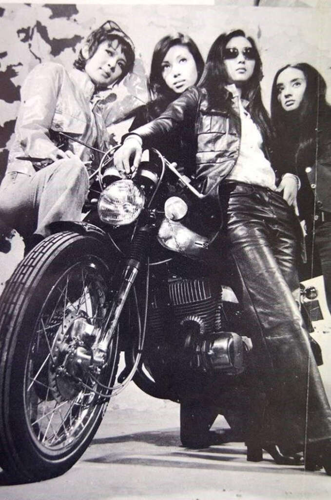 Four individuals pose confidently around a motorcycle in this black-and-white photo. The two in front wear leather jackets and sunglasses, exuding a cool, rebellious vibe. The other two are standing behind them, completing a stylish, bold group ensemble.