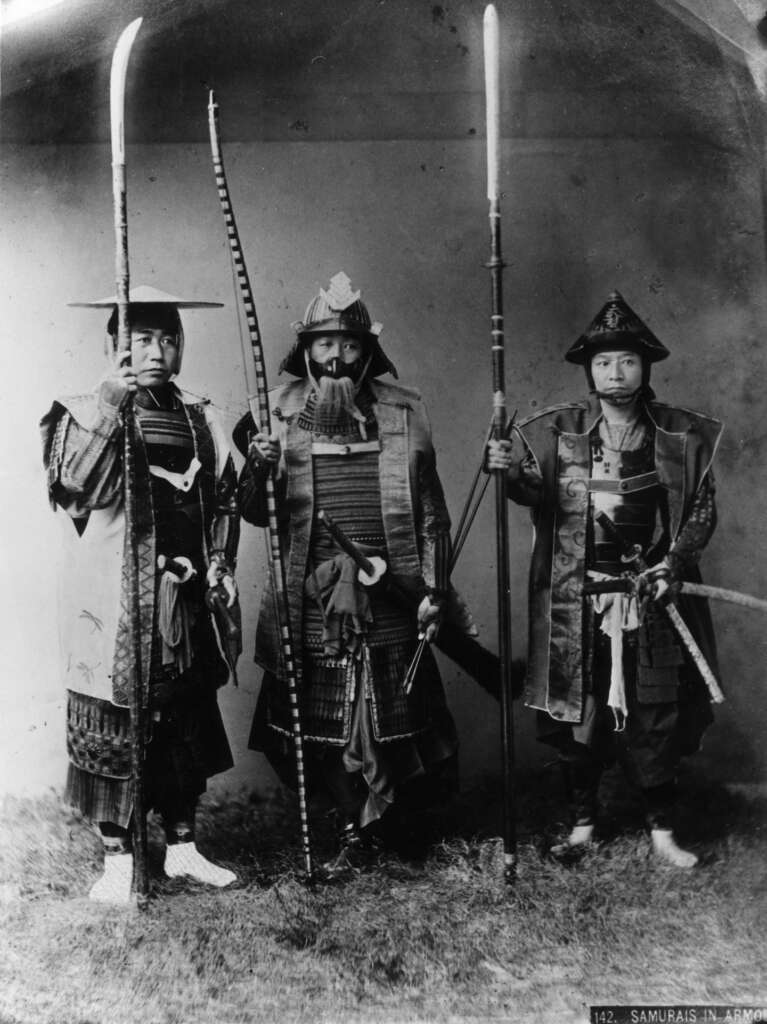 Three samurais in traditional armor stand together, each holding long weapons. The photo is black and white, showcasing detailed armor and helmets, with the warriors looking directly at the camera.