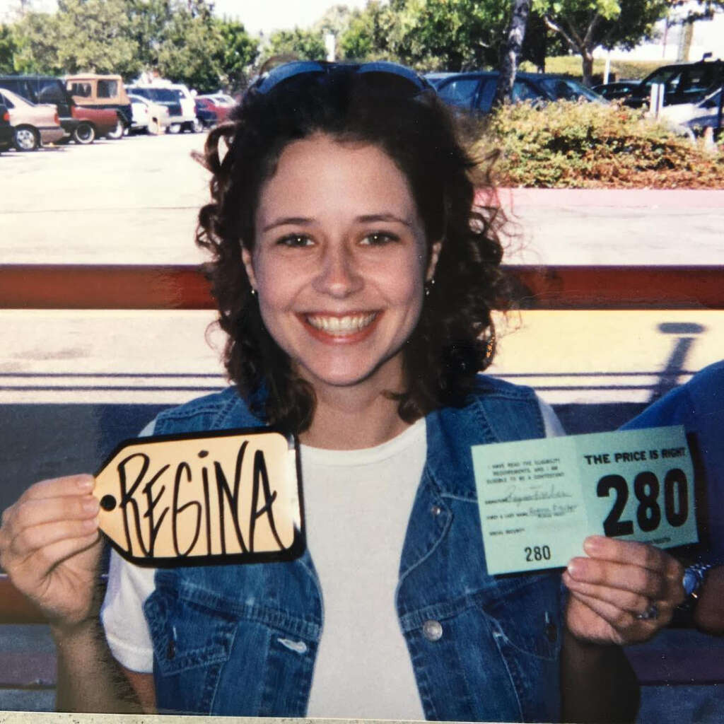 A woman smiling broadly holds a card with "Regina" written on it and another card with "The Price Is Right 280" in her hands. She is wearing a white shirt and a denim vest. Trees and cars are visible in the background.