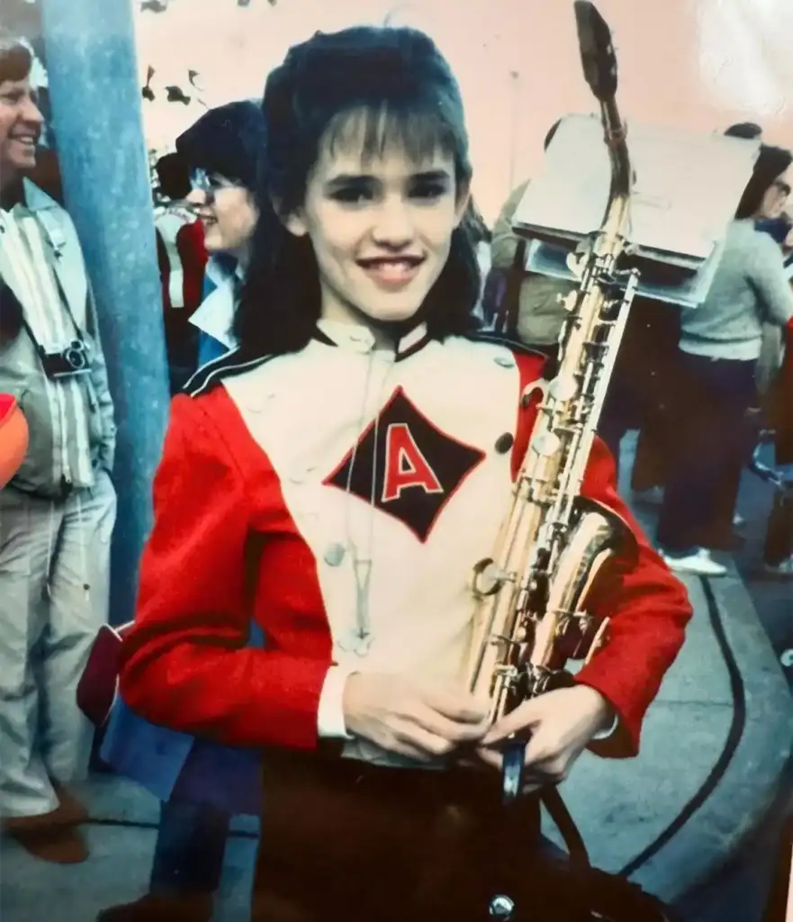 A young woman in a red and white marching band uniform holds a saxophone. She is smiling at the camera. People with cameras and casual attire are gathered in the background.