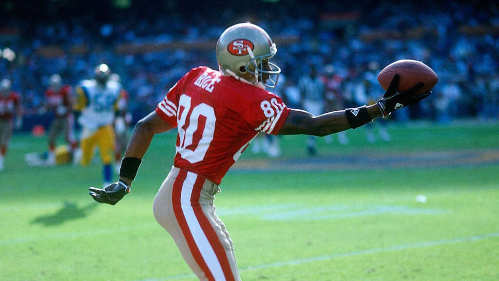 A football player in a red jersey with the number 80 catches a ball with one hand during a game. He is wearing a helmet and gloves, and the sunny field is visible in the background. Players from the opposing team are seen in the distance.