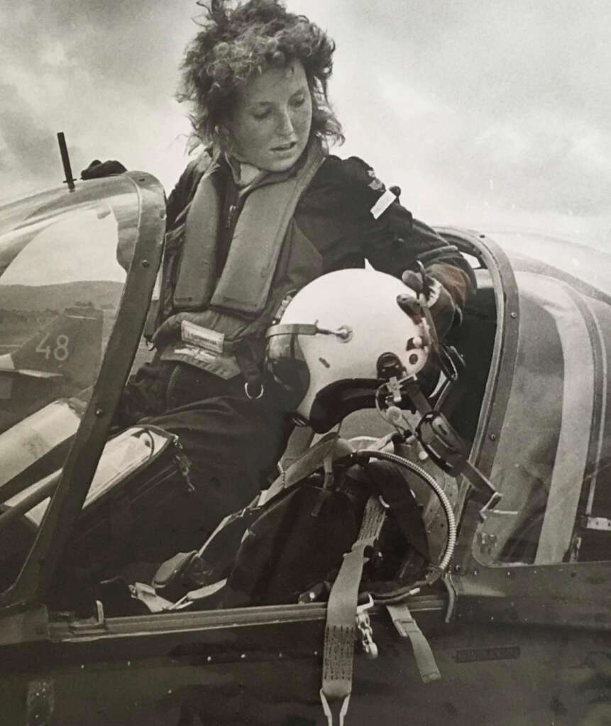 A person with curly hair, wearing a flight suit and life vest, stands by the cockpit of a small aircraft, holding a white helmet. The sky is cloudy in the background.