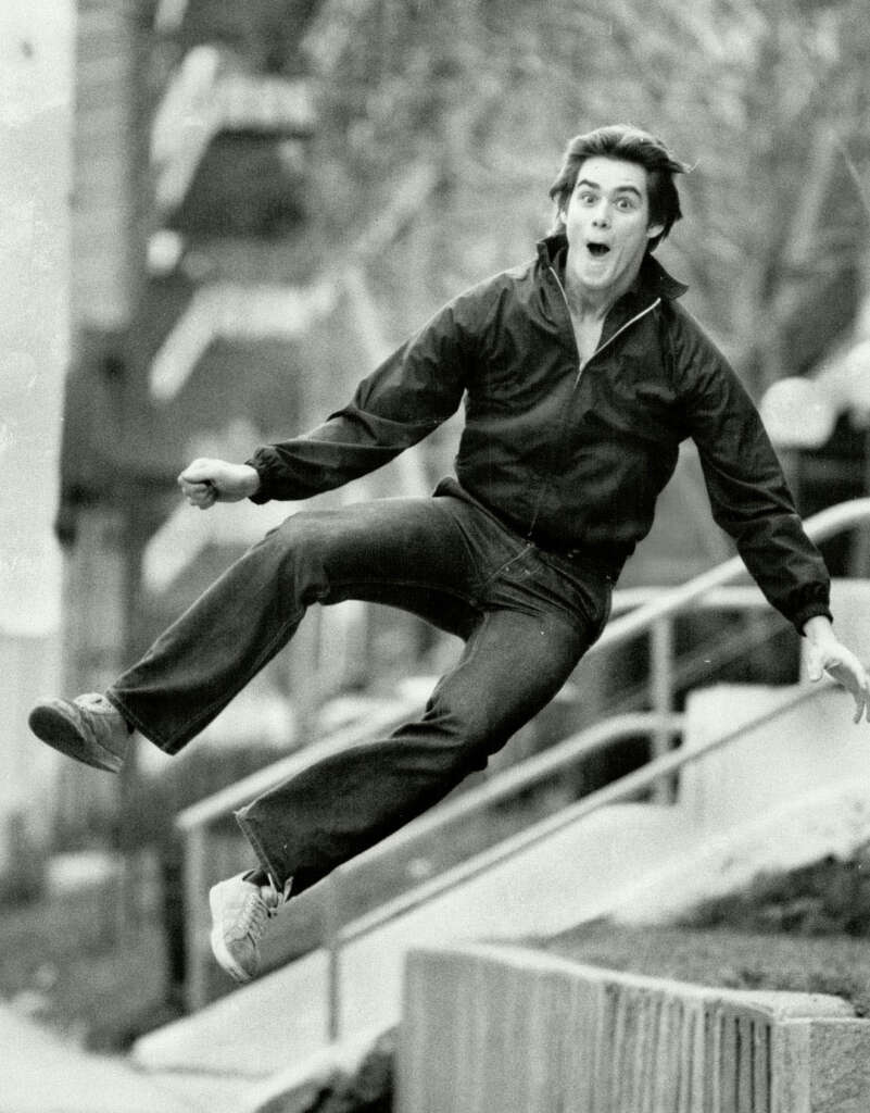 A person in a dark jacket and jeans is captured mid-air while jumping. They have an expressive, surprised look on their face. The background includes a blurred outdoor setting with a railing and trees.