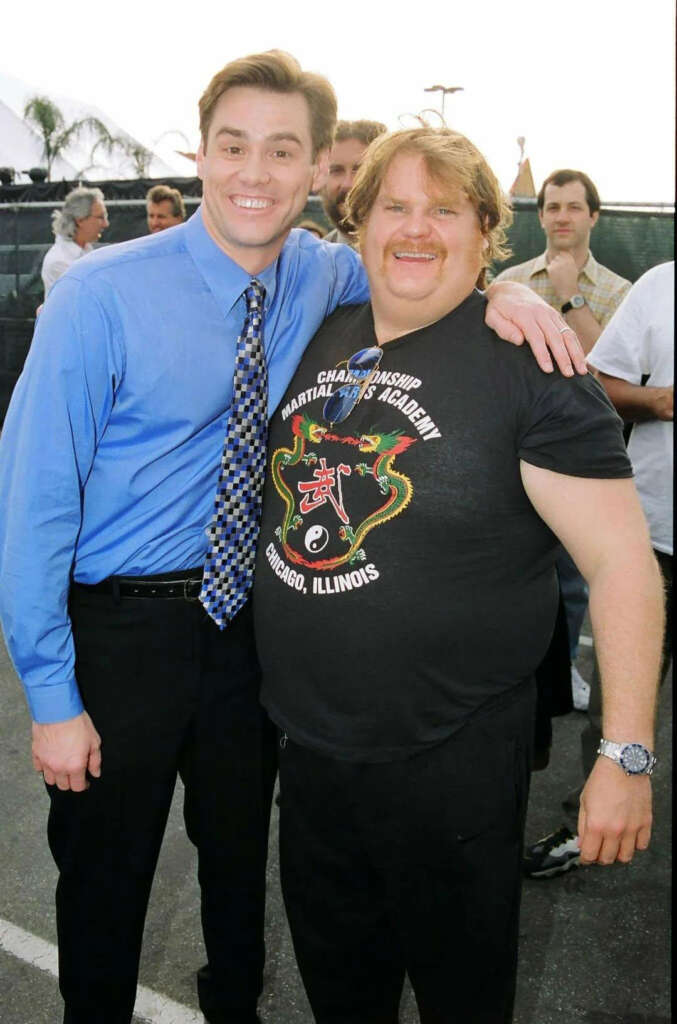 Two people are smiling for the camera. The person on the left is wearing a blue shirt and patterned tie, while the person on the right is in a black martial arts t-shirt. They are standing outdoors with others in the background.