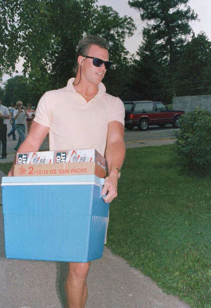 A person wearing sunglasses carries a blue cooler and a box labeled "2-12/12 OZ CAN PACKS" outdoors. A red SUV is parked in the background, with trees and grass nearby.